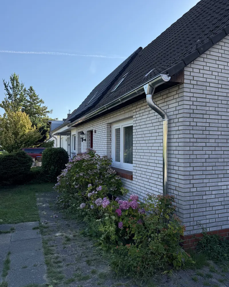 Hausseite mit neuer Dachrinne und Fallrohr, davor blühende Sträucher