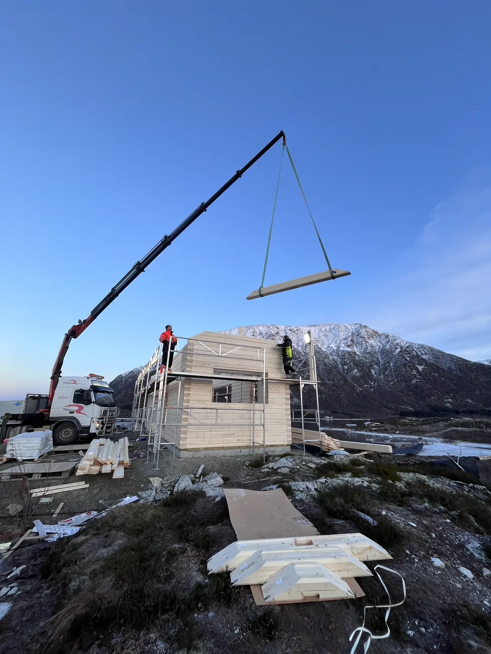 Kran hebt einen vorgefertigten Dachbinder vor winterlichem Bergpanorama